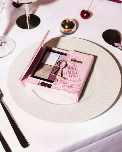 Růžova paleta očnich stinů Lancome se zlatym motivem kličove dirky, elegantně umistěna na bilem jidelnim taliři.