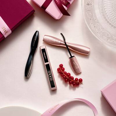 Pohled shora na produkty dekorativni kosmetiky Lancome - řasenku a lesk na rty - doplněne růžovymi darkovymi krabičkami, satenovymi stuhami a snitkou červeneho rybizu.