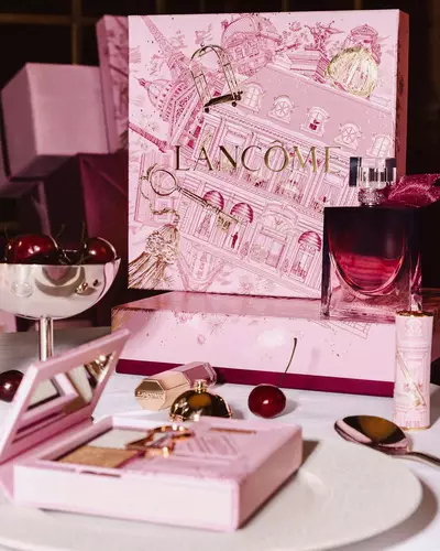 Svatečni expozice Lancome s velkou ilustrovanou růžovou darkovou krabičkou, parfemem a produkty dekorativni kosmetiky naaranžovanymi na stole s čerstvymi třešněmi.