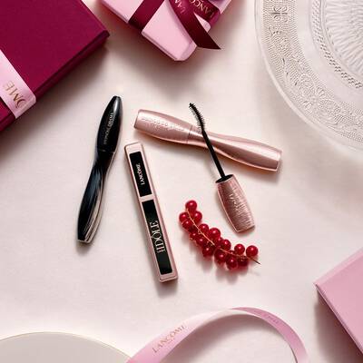 Kompozice make-upovych produktů Lancome focenych shora, včetně řasenky a lesku na rty, spolu s růžovymi darkovymi krabičkami, satenovymi stuhami a snitkou červeneho rybizu.