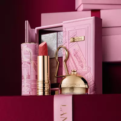 Pečlivě sestavena svatečni darkova sada Lancome s rtěnkou, paletou očnich stinů a zlatymi kosmetickymi produkty vystavenymi na dekorativnim růžovem pouzdře.