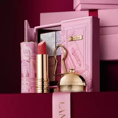Pečlivě naaranžovana svatečni darkova kolekce Lancome s rtěnkou, paletkou očnich stinů a zlatymi kosmetickymi produkty vystavenymi na dekorativnim růžovem pouzdře.