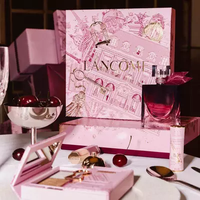 Svatečni kolekce Lancome naaranžovana na stole, včetně velke růžove darkove krabičky s ilustracemi, parfemu, kosmetickych produktů a čerstvych třešni.