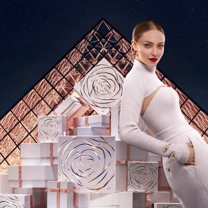 Žena v elegantnim bilem oblečeni sedi vedle hromady darkovych krabiček před skleněnou pyramidou muzea Louvre.