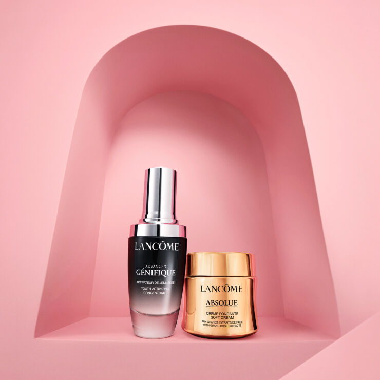 Na obrazku jsou dva kosmeticke produkty značky Lancome: serum Advanced Genifique a krem Absolue, umistěne na růžovem pozadi.