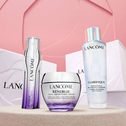 Na obrazku jsou tři produkty značka Lancome: serum, anti-aging krem a dvojita esence, umistěne na jemně růžovem pozadi s dekorativnimi boxy.