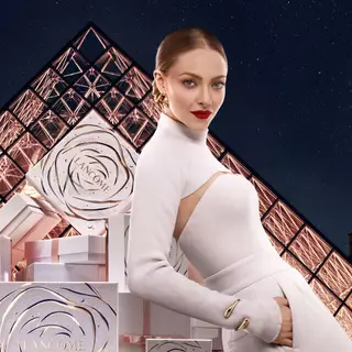 Modelka v bilem oblečeni s červenymi rtěnkami, stojici před darkovymi balenimi Lancome a pozadim s pyramidou.
