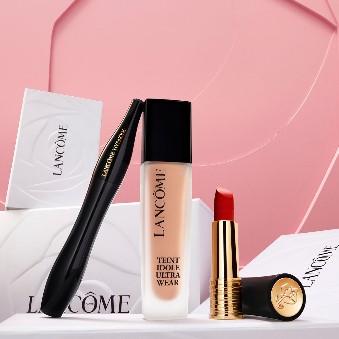 Na obrazku je ruka, ktera ukazuje na kosmeticke produkty značky Lancome, jako je make-up a rtěnka, umistěne na bilych krabičkach na růžovem pozadi.