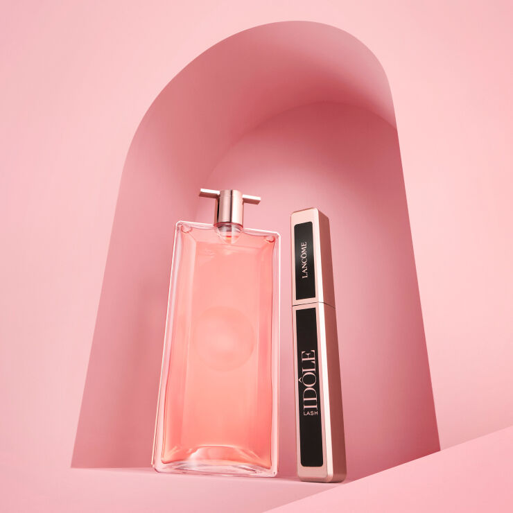 Na obrazku je růžova parfemova flakon a řasenka, umistěne na pozadi v pastelově růžove barvě.