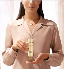 A woman in a beige blouse holds a bottle of Lancome skincare product, displaying it prominently in front of her.