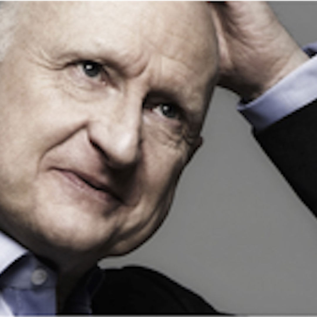 A middle-aged man in a dark suit and light blue shirt is thoughtfully touching his head with a slight smile, against a neutral background.