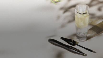 Na obrazku je kosmeticky produkt Lancome, zahrnujici čistici a pečujici prostředek v průhledne lahvičce a řasenku. V pozadi jsou jemne rostlinne motivy.