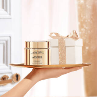 A hand holds a golden tray with a jar of Lancome Absolue cream and a beautifully wrapped gift box against a softly lit background with shimmering curtains.