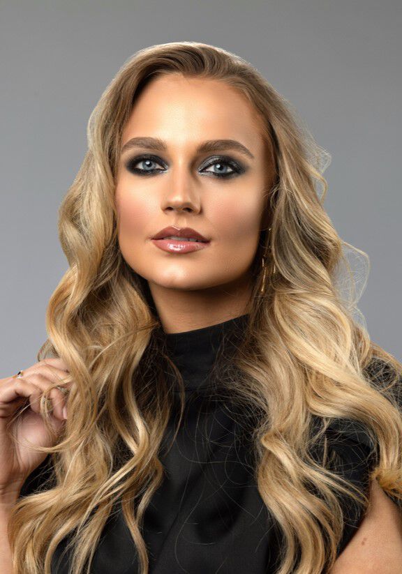 Žena s dlouhymi vlnitymi vlasy a vyraznym makeupem, oblečena v černem topu, stoji před neutralnim pozadim.