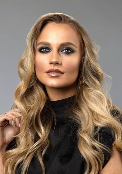 Žena s dlouhymi vlnitymi vlasy a vyraznym makeupem, oblečena v černem topu, stoji před neutralnim pozadim.
