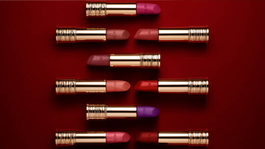 A variety of lipsticks with gold packaging arranged in a grid pattern on a red background, showcasing different shades of pink, purple, and coral.