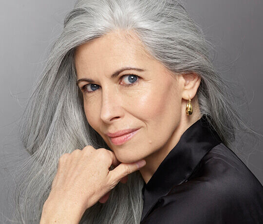A close-up portrait of an elegant woman with long, flowing gray hair, posing with her chin resting on her hand. She has striking blue eyes and subtle makeup, wearing a black blouse and simple gold earrings.