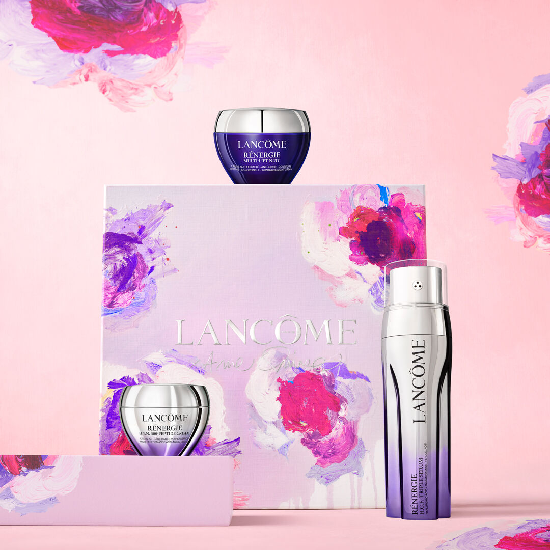 Alt text: A product display featuring Lancome skincare items, including two jars and a bottle, on a pastel pink background with colorful floral accents. The packaging is beautifully designed with the Lancome logo.