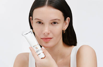 A woman with long dark hair holds a skincare device while smiling at the camera against a plain white background.