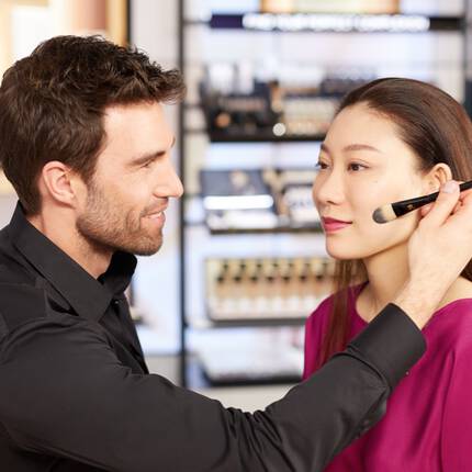A makeup artist applies foundation to a woman's face using a brush, while she sits in a well-lit beauty store filled with makeup products in the background.