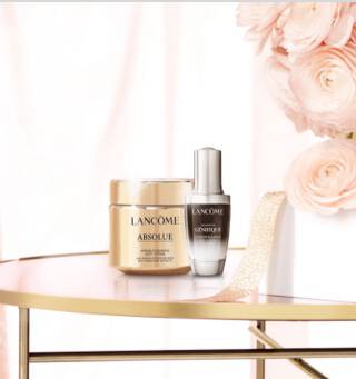 A luxurious display featuring Lancome skincare products, including a gold jar of Absolue cream and a Genifique serum, alongside a bouquet of soft pink flowers on a stylish table.