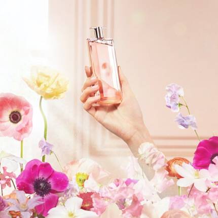 A hand holds a bottle of perfume amidst a blooming garden of colorful flowers, with soft, light-filtering through a window in the background.