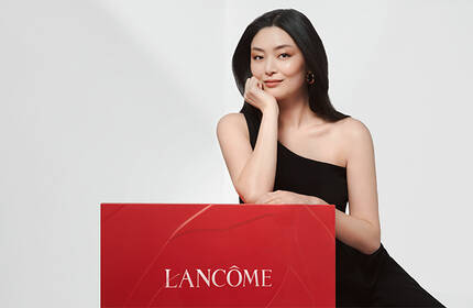 A woman in a black dress is posing next to a large red box with the Lancome logo on it, set against a light background.