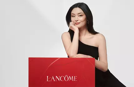 A woman in a black dress is posing next to a large red box with the Lancome logo on it, set against a light background.
