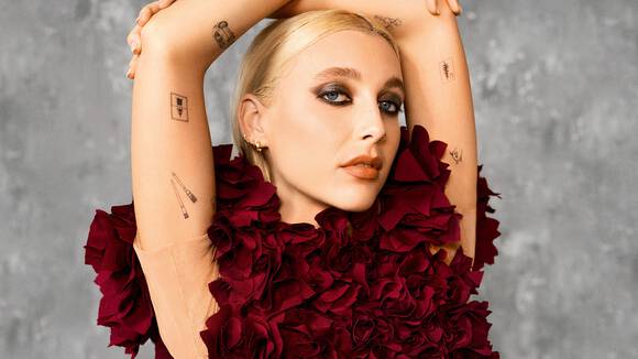 A person with blonde hair styled back is posing with their arms raised, showcasing a dress adorned with large burgundy flower petals. Their makeup features bold eye makeup, and they have various small tattoos on their arms. The background is a neutral gray.
