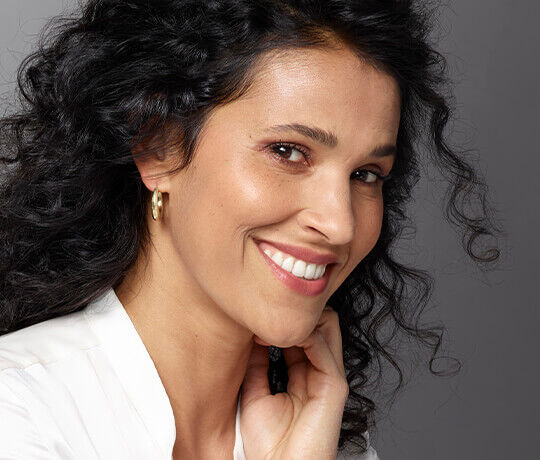A smiling woman with curly black hair, wearing hoop earrings, rests her chin on her hand against a neutral background.