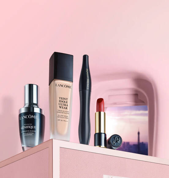 A collection of Lancome beauty products displayed on a pink surface, including a serum, foundation, mascara, and red lipstick.