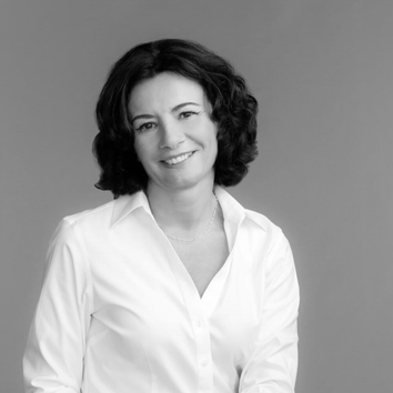 Černobily portret ženy s kudrnatymi vlasy, oblečene v bile košili, usmivajici se do kamery.