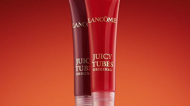 Two lip gloss tubes from Lancome's Juicy Tubes collection, one in a deep red shade and the other in a brighter red, set against a warm gradient background.
