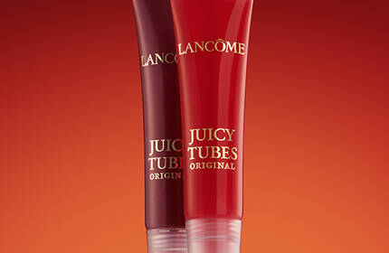 Two lip gloss tubes from Lancome's Juicy Tubes collection, one in a deep red shade and the other in a brighter red, set against a warm gradient background.