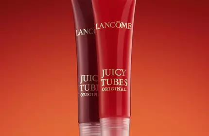 Two lip gloss tubes from Lancome's Juicy Tubes collection, one in a deep red shade and the other in a brighter red, set against a warm gradient background.