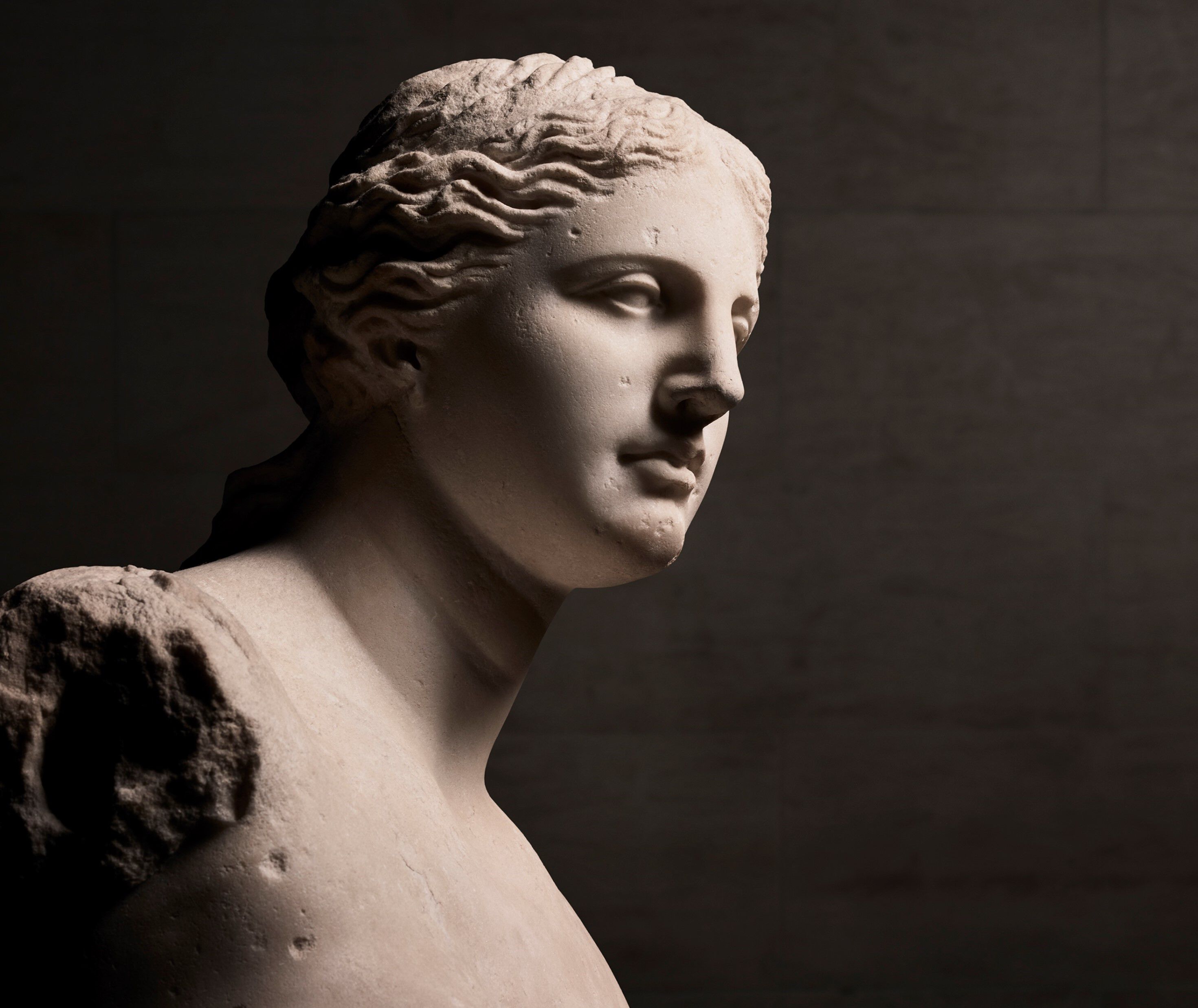 A close-up view of a marble statue of a woman's head and shoulders, featuring classical details and softly illuminated to emphasize its texture and form.