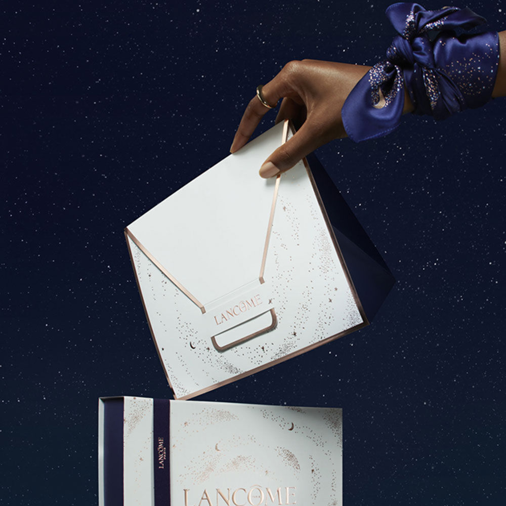 A hand elegantly holds a white envelope with gold accents labeled "Lancome," above a beautifully designed gift box set against a starry night background.