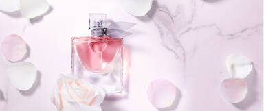 A bottle of pink perfume with a silver ribbon, surrounded by white rose petals on a marble surface.