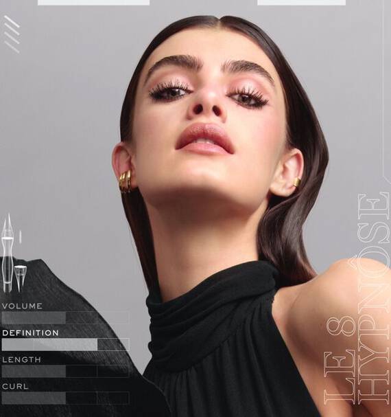 A close-up portrait of a model with long, straight hair wearing a sleeveless black top. She poses confidently, gazing upwards with a bold makeup look, featuring dramatic eyelashes and glossy lips. The image includes text about mascara features: volume, definition, length, and curl.