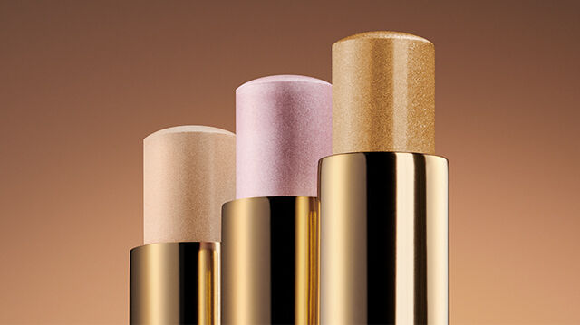 Three makeup sticks in gold packaging arranged vertically on a gradient brown background. The sticks feature varying shades: light beige, soft pink, and a shimmer gold.
