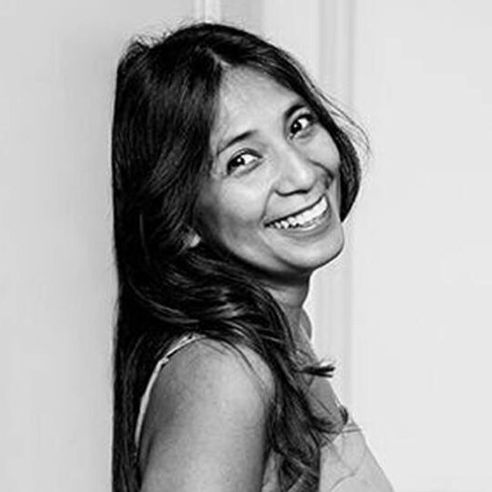 A woman with long, dark hair smiles brightly while leaning against a wall in a black and white photograph.
