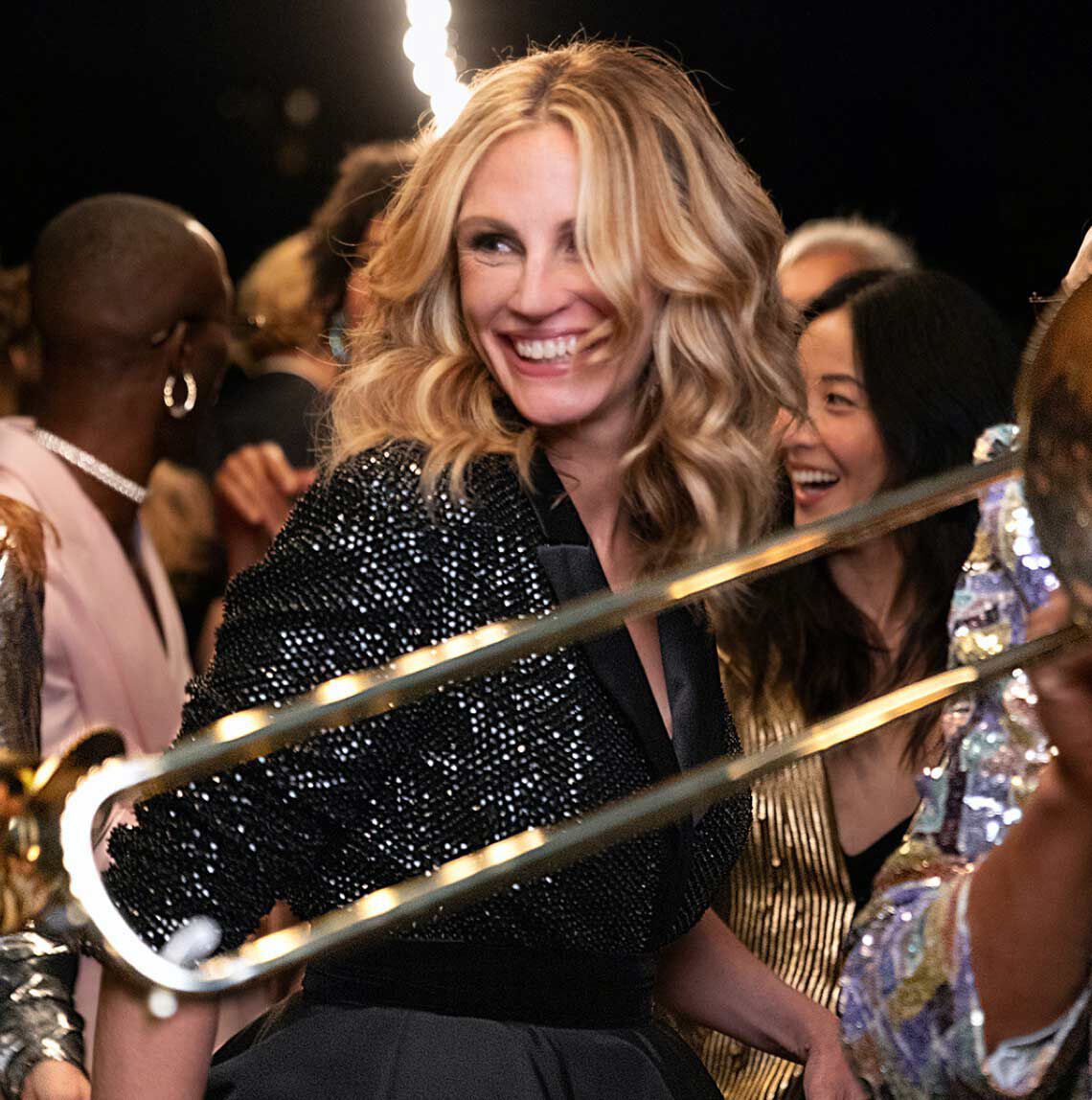 Žena s vlasy po ramena, oblečena ve třpytivem saku, se usmiva uprostřed oslavy, obklopena lidmi a hudebnimi nastroji.