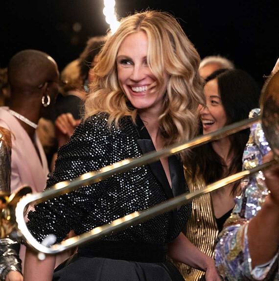 Žena s vlasy po ramena, oblečena ve třpytivem saku, se usmiva uprostřed oslavy, obklopena lidmi a hudebnimi nastroji.