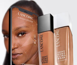 Na obrazku je kosmeticky produkt značky Lancome, konkretně make-up "Teint Idole Ultra Wear," se zobrazenim jeho lahvičky vedle mobilniho telefonu, na jehož obrazovce je vyobrazena modelka.