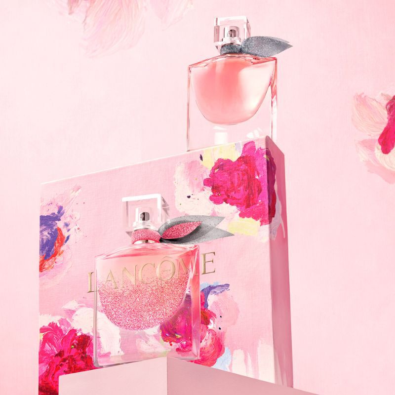 Two elegant perfume bottles are displayed atop a beautifully decorated box featuring vibrant pink and floral designs. The background is a soft pink, enhancing the overall romantic aesthetic.