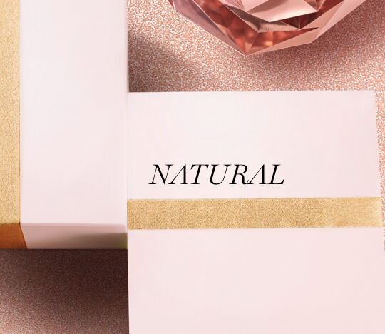 A close-up of two elegantly wrapped gift boxes with a soft pink hue and a gold ribbon, featuring the word "NATURAL" in bold black text. In the background, there's a decorative rose-colored crystal.