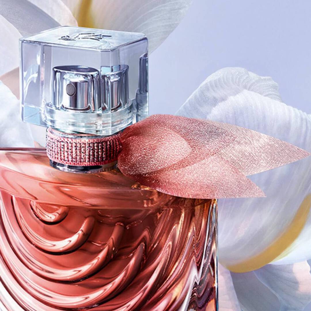 A close-up of a stylish perfume bottle with a textured coral-colored liquid, featuring a clear cap and a pink ribbon accent, set against a soft floral background.