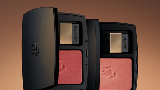 A close-up of two elegant black compact cases containing pink blushes, set against a warm, gradient background.