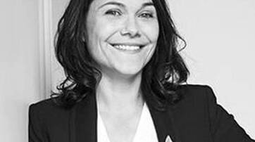 A smiling woman with shoulder-length dark hair wearing a blazer, posing confidently against a plain background.