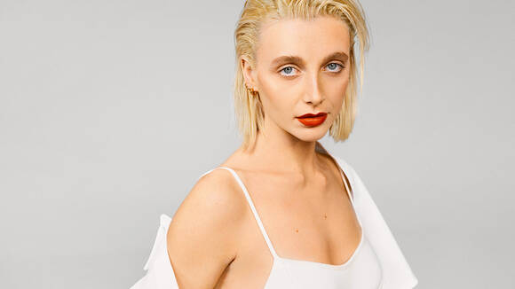 A person with short blonde hair and striking blue eyes poses confidently, wearing a white dress and bold red lipstick, against a neutral gray background.